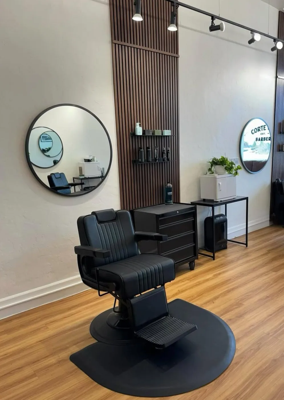 A single black leather barber chair beneath a round black-framed mirror and walnut slat wall
